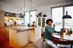 Smiling male sitting at his dining table with his laptop. Kitchen in the background.

