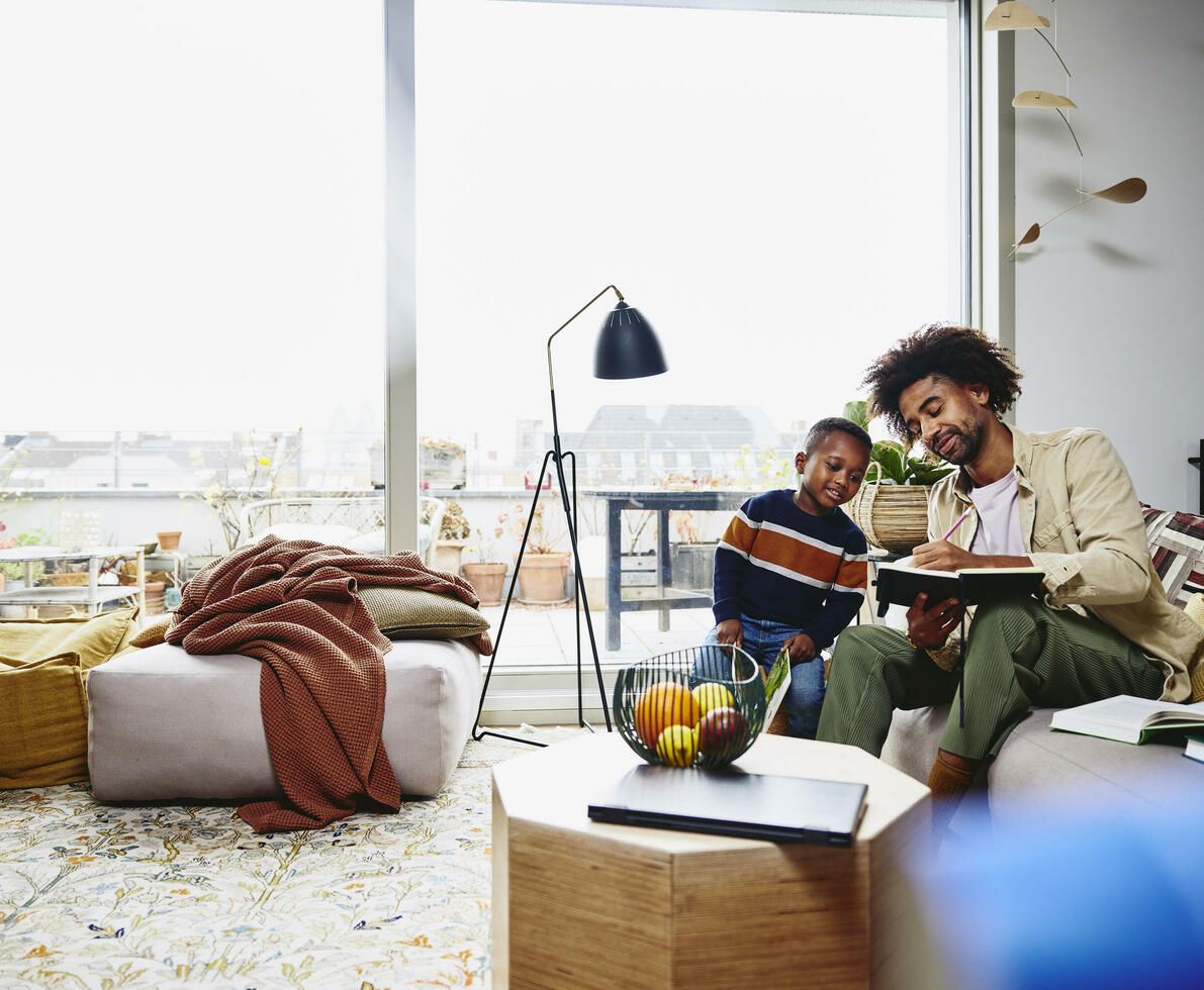 male sitting on couch holding a notebook and pen. Showing his notebook to a kid.
